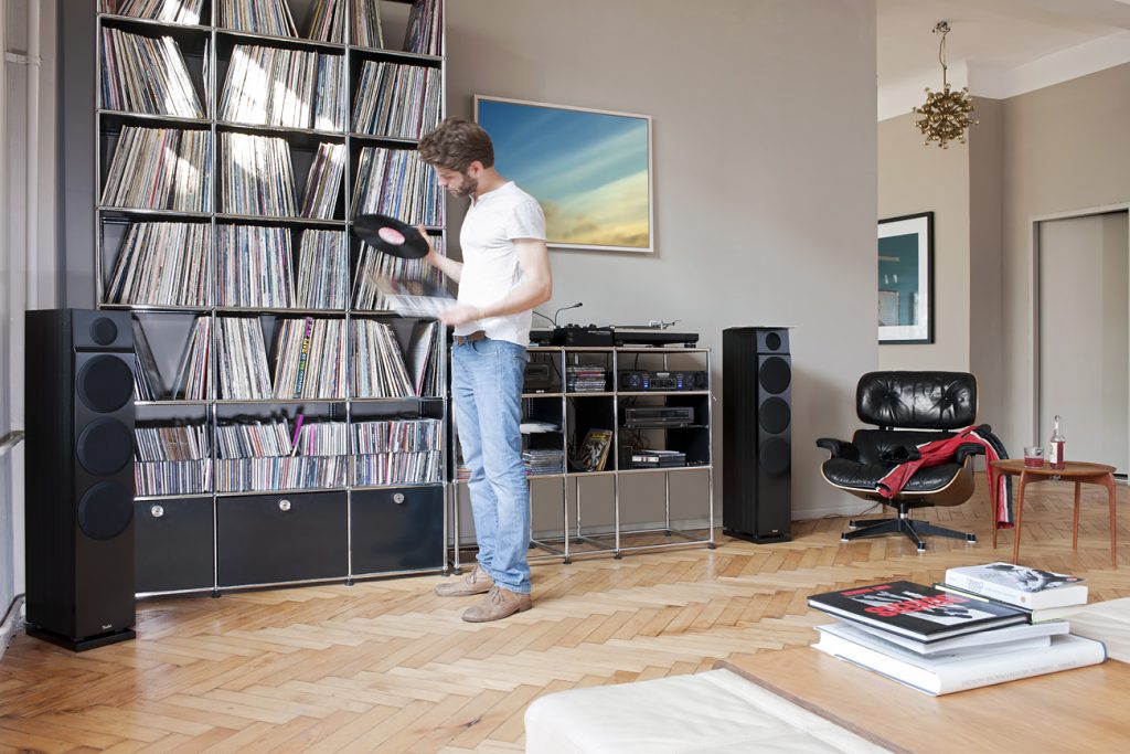 Ein Mann steht in einem Wohnzimmer und schaut auf eine Schallplatte, die er in der Hand hält.
