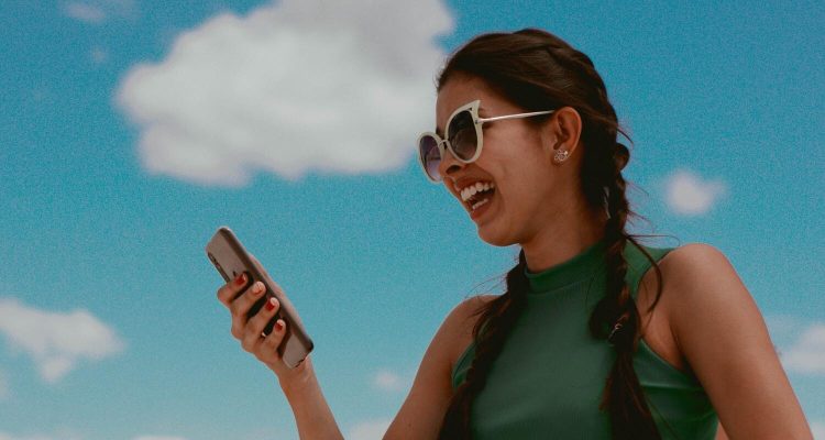 Eine junge Frau lacht und hält das Handy vor sich. Im Hintergrund ist blauer Himmel zu sehen.