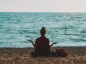 Eine Frau meditiert am Strand, im Hintergrund ist das ruhige Meer zu sehen.