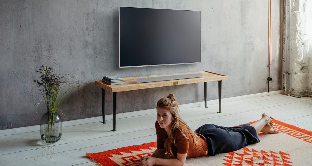 Frau liegt im Wohnzimmer auf dem Boden, im Hintergrund sieht man einen an der Wand hängenden Smart-TV und eine Soundbar. 