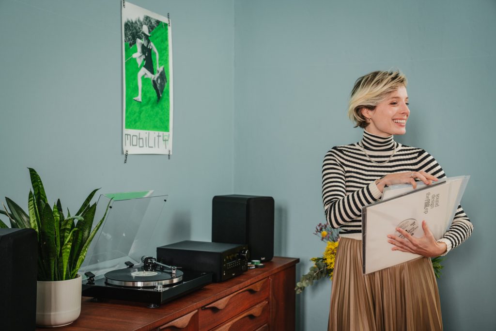 Frau mit Schallplatten im Arm vor einer Komode mit Plattenspieler und Lautsprecher.