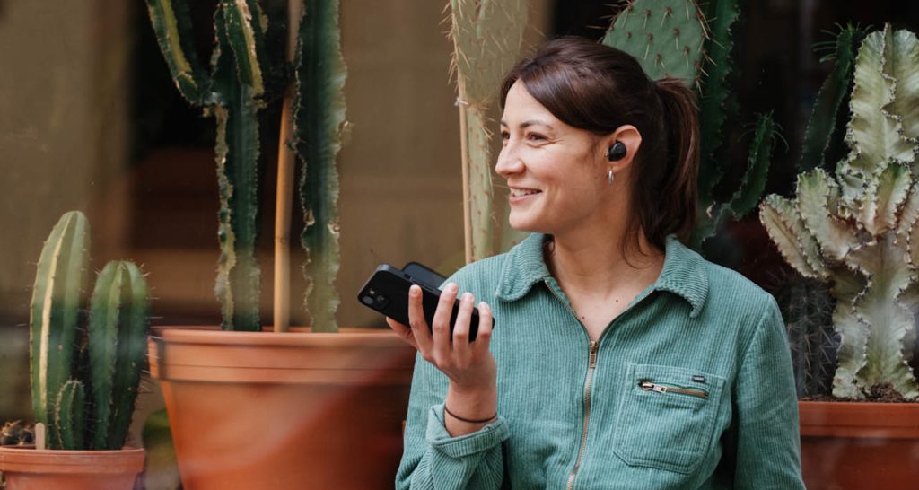 Eine lächelnde Frau trägt In-Ear-Kopfhörer und hält ein Smartphone in der Hand.