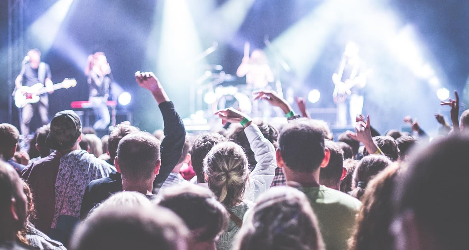 Publikum mit erhobenen Armen vor einer hell beleuchteten Konzertbühne, auf der eine Band live spielt