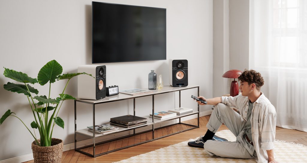 homme assis sur son tapis devant sa télé avec les enceintes ultima 25 active s'apprête à regarder une série dans son salon ,la télécommande à la main 