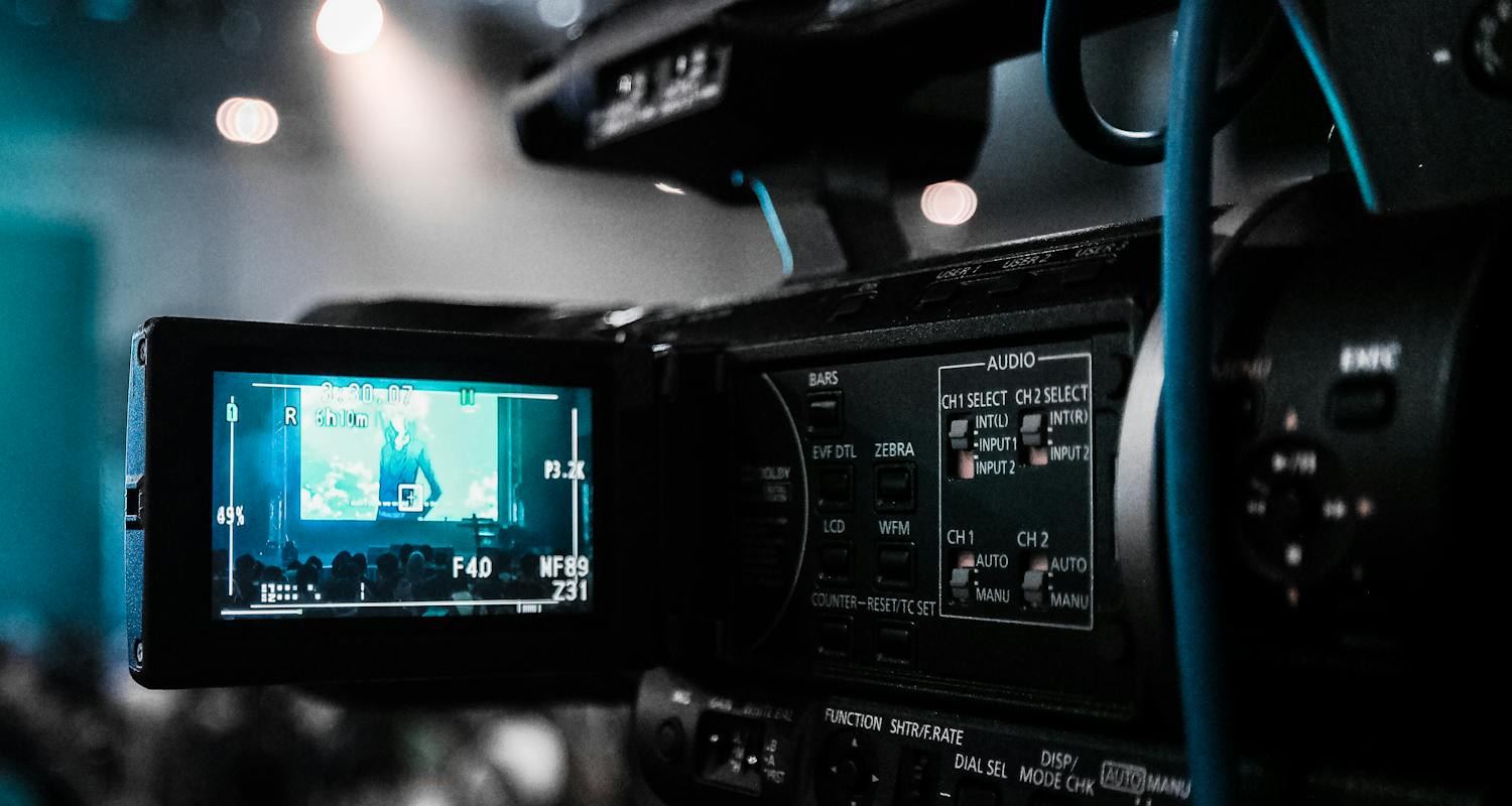 Nahaufnahme einer Videokamera, die eine Veranstaltung mit Publikum und Bühne filmt, sichtbar im Kameradisplay.