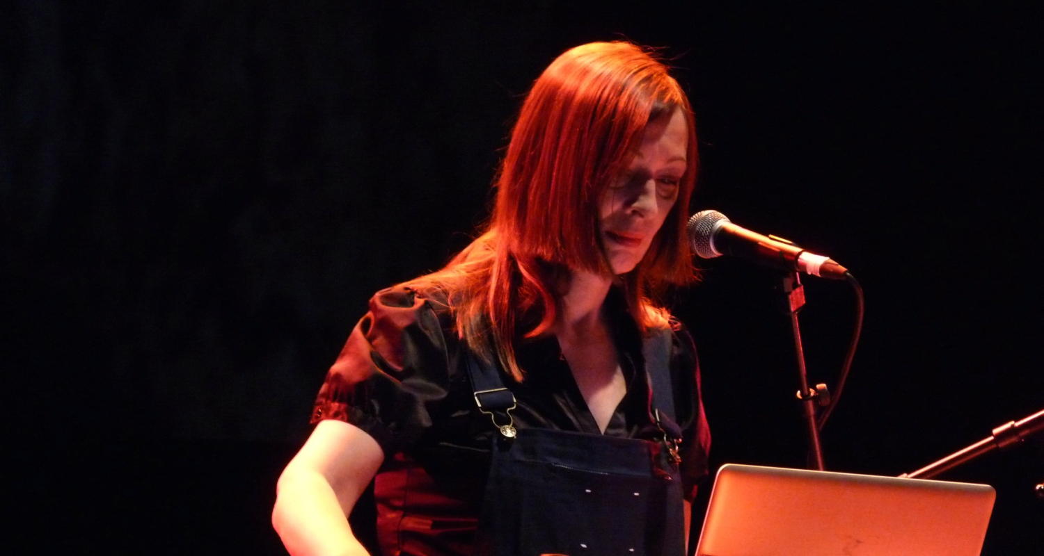 Gudrun Gut live auf der Bühne mit Laptop und Mikrofon bei einer elektronischen Performance im roten Bühnenlicht.