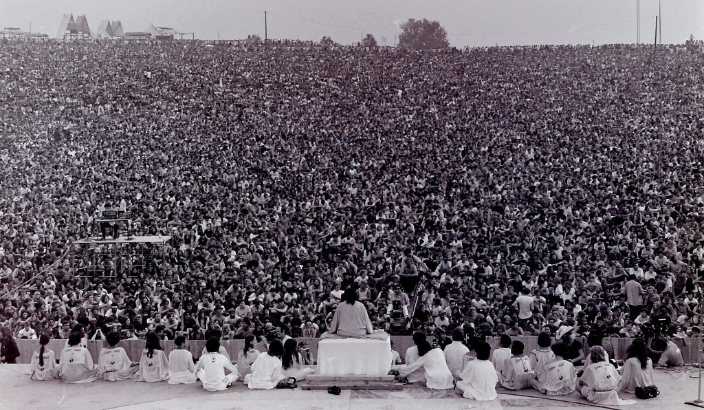 Swami Satchidananda und seine Begleiter sitzen bei der Eröffnungszeremonie von Woodstock 1969 auf der Bühne vor einer riesigen Menschenmenge.